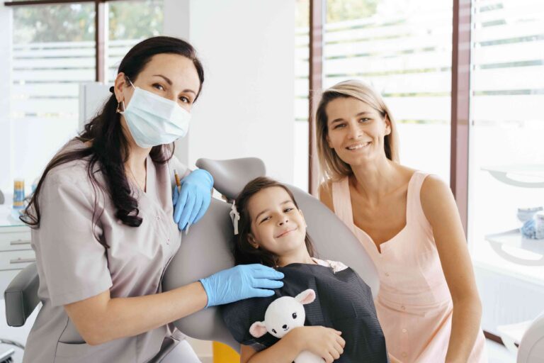 Dentist providing a child-friendly dental exam while a mother sits beside her smiling daughter.
