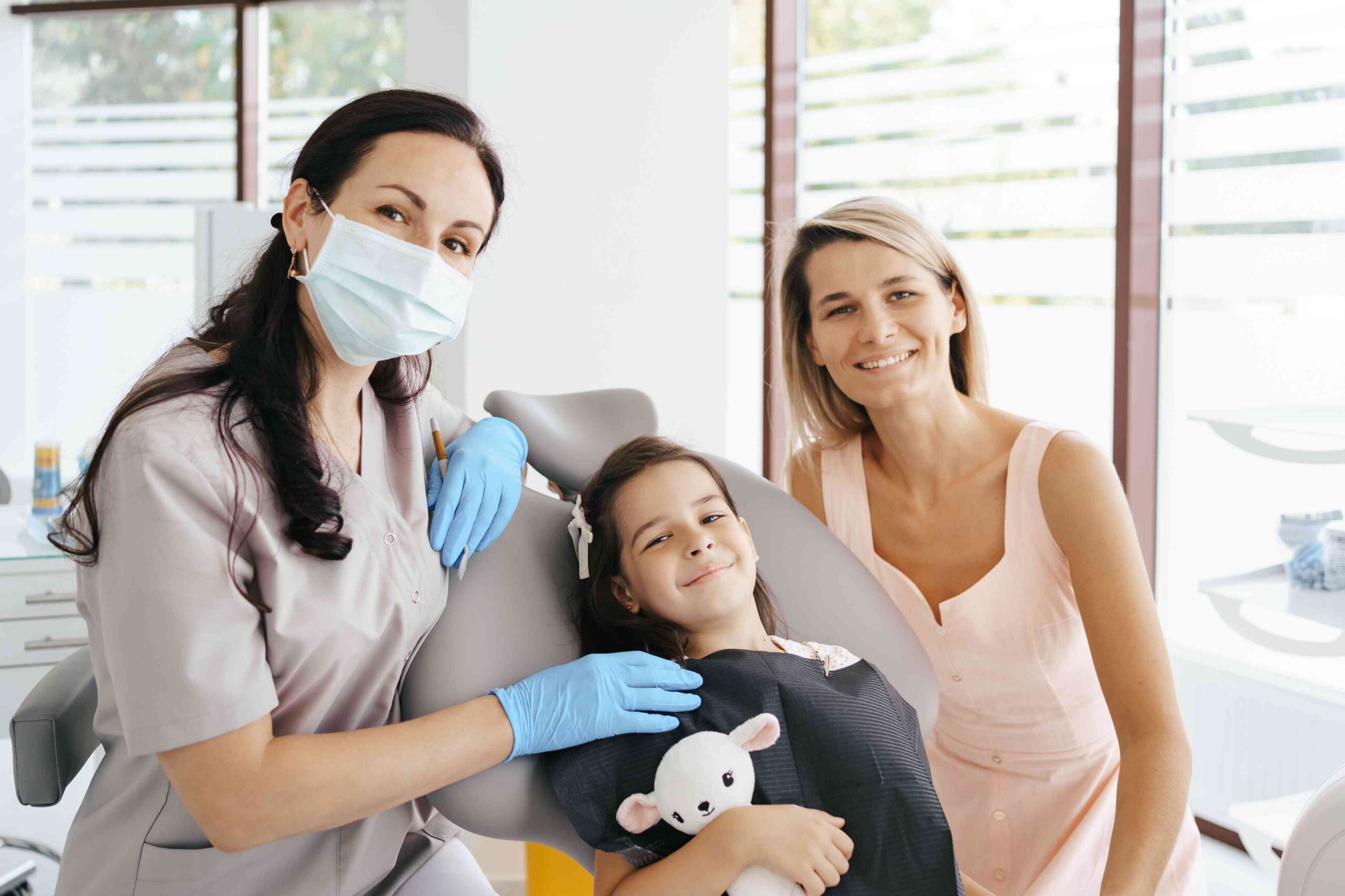 Dentist providing a child-friendly dental exam while a mother sits beside her smiling daughter.